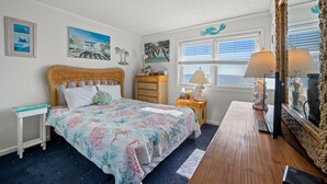 A bedroom with a bed, a wooden nightstand, a wicker chair, a wicker dresser, a mirror, and a window with blinds.