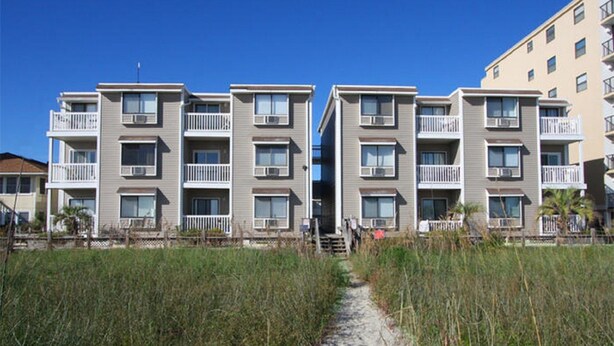 Un edificio de apartamentos frente a la playa con varios balcones, un camino de arena que conduce a la entrada y un cielo azul despejado.
