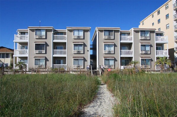 A beachfront apartment building with multiple balconies, a sandy pathway leading to the entrance, and a clear blue sky.