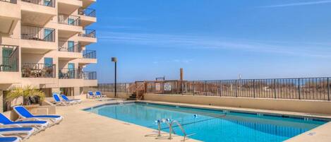 A swimming pool with blue lounge chairs, a concrete deck, and a building with balconies.