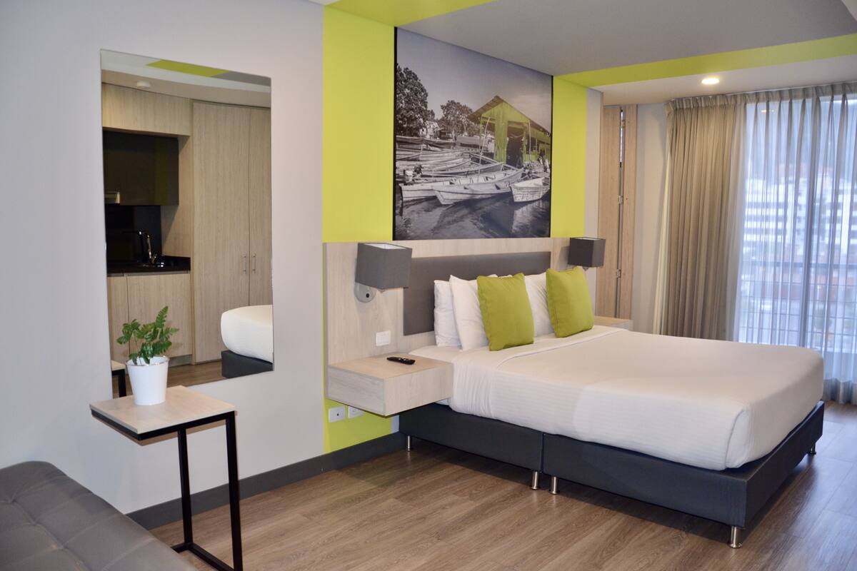 A modern hotel room with a large bed, bedside tables, a small desk, a mirror, and a painting on the wall.