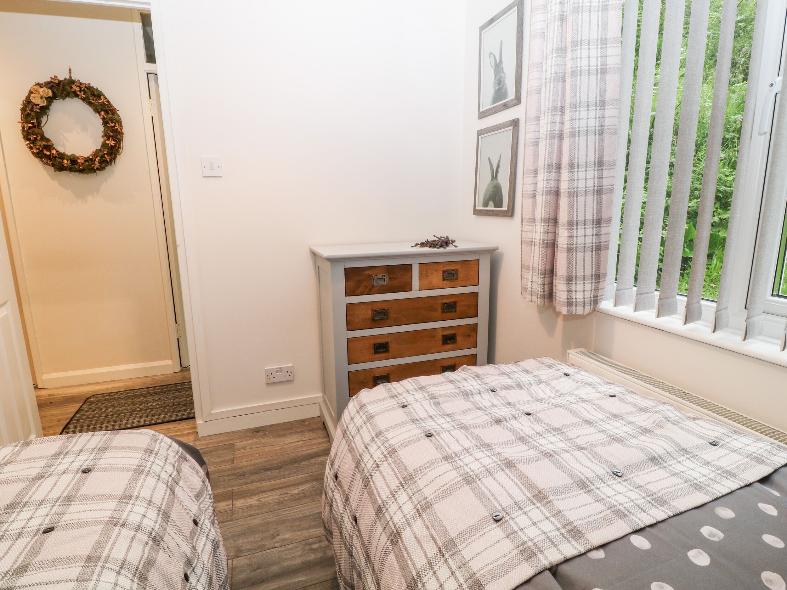 A bedroom with two beds, a dresser, a window with curtains, and a wall-mounted wreath.