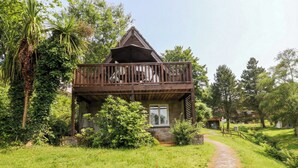 A two-story wooden house with a balcony, surrounded by lush greenery and a gravel path leading to the entrance. - Lavender Lodge (Gunnislake)