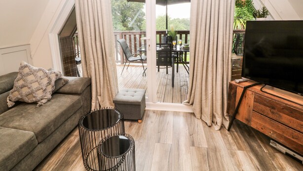 A cozy living room with a sofa, a TV, and a wooden floor. There is a balcony with a table and chairs, and a large window with curtains. - Lavender Lodge (Gunnislake)
