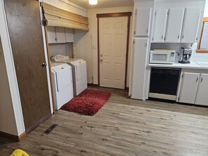 Cocina compacta con microondas, cafetera y lavavajillas. Hay un tapete rojo en el suelo y una puerta de madera.