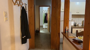 A narrow hallway with wooden walls, a coat rack, and a glimpse of a kitchen area with cabinets and a countertop. - Cosy Calm Cottage in Olive Trees With sea View (Ermionida)