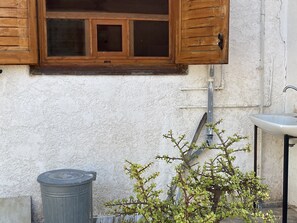 A window with wooden shutters, a white wall, a small plant, a trash bin, and a sink. - Cosy Calm Cottage in Olive Trees With sea View (Ermionida)