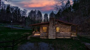 Una cabaña rústica de troncos con chimenea de piedra, un porche con asientos y vistas al bosque circundante.