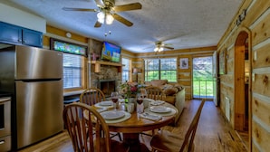 Interior de una cabaña de madera con comedor, cocina y un gran ventanal con vistas al exterior.