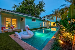 Une piscine avec un flotteur en forme de cygne, un patio couvert avec des chaises et une maison à l’extérieur bleu clair.