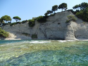 Paisagem costeira com águas cristalinas, uma praia de areia e um penhasco rochoso com árvores.