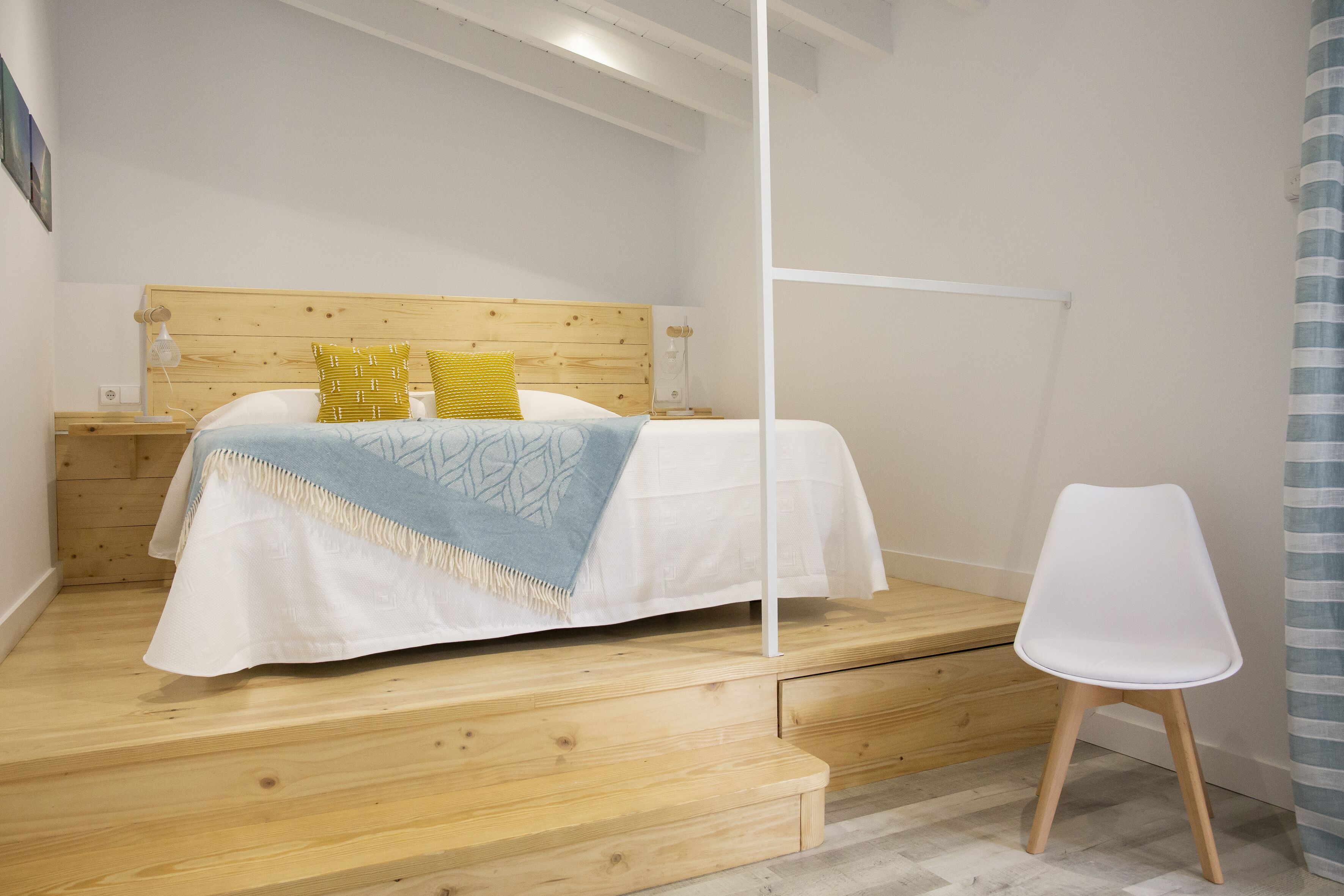Un dormitorio con cabecera de madera, cama con ropa de cama blanca y una manta azul, una silla blanca y una mesita de noche de madera con una lámpara.