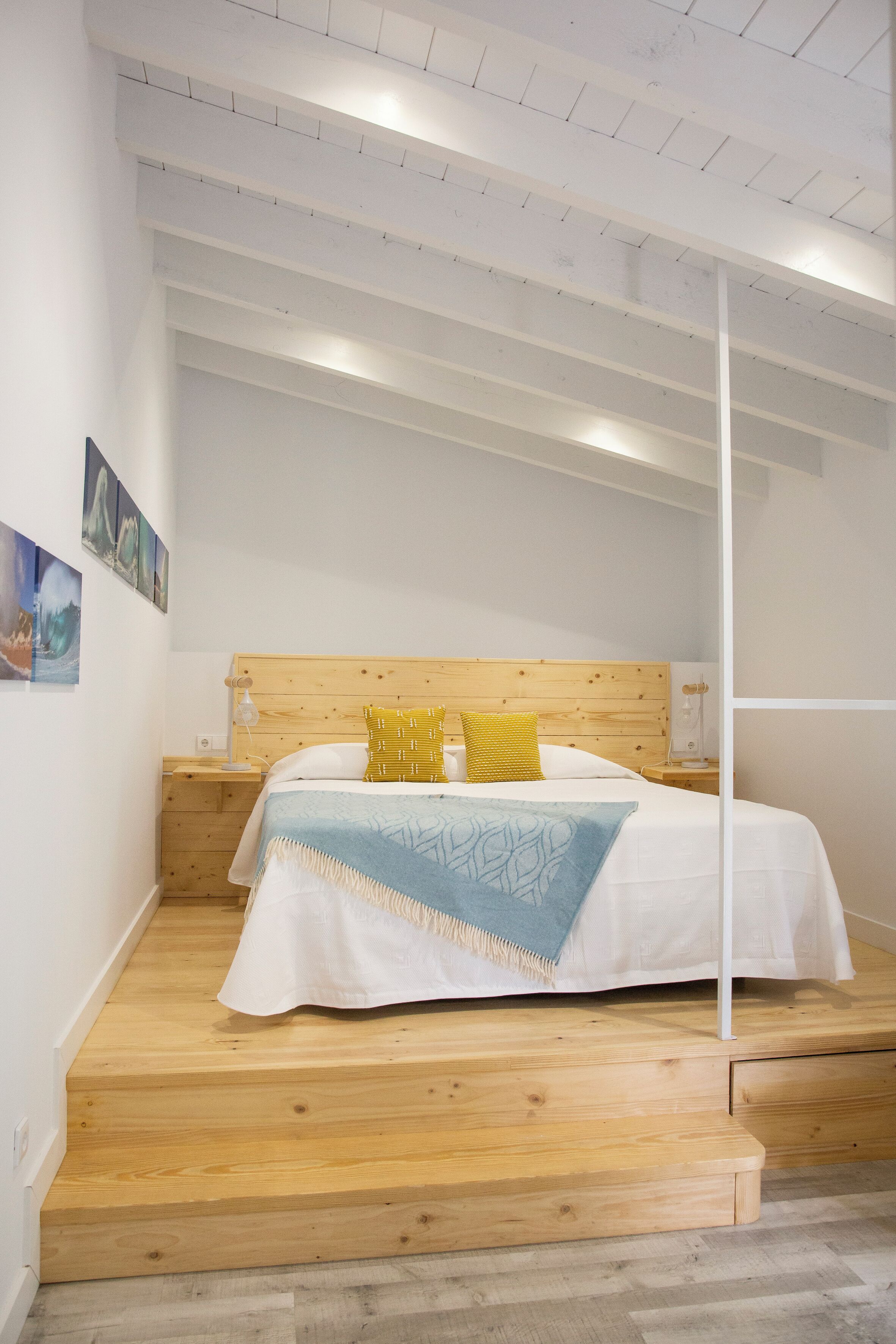 A bedroom with a wooden headboard, a bed with white bedding and a blue throw, a wooden nightstand, and a wall-mounted shelf.
