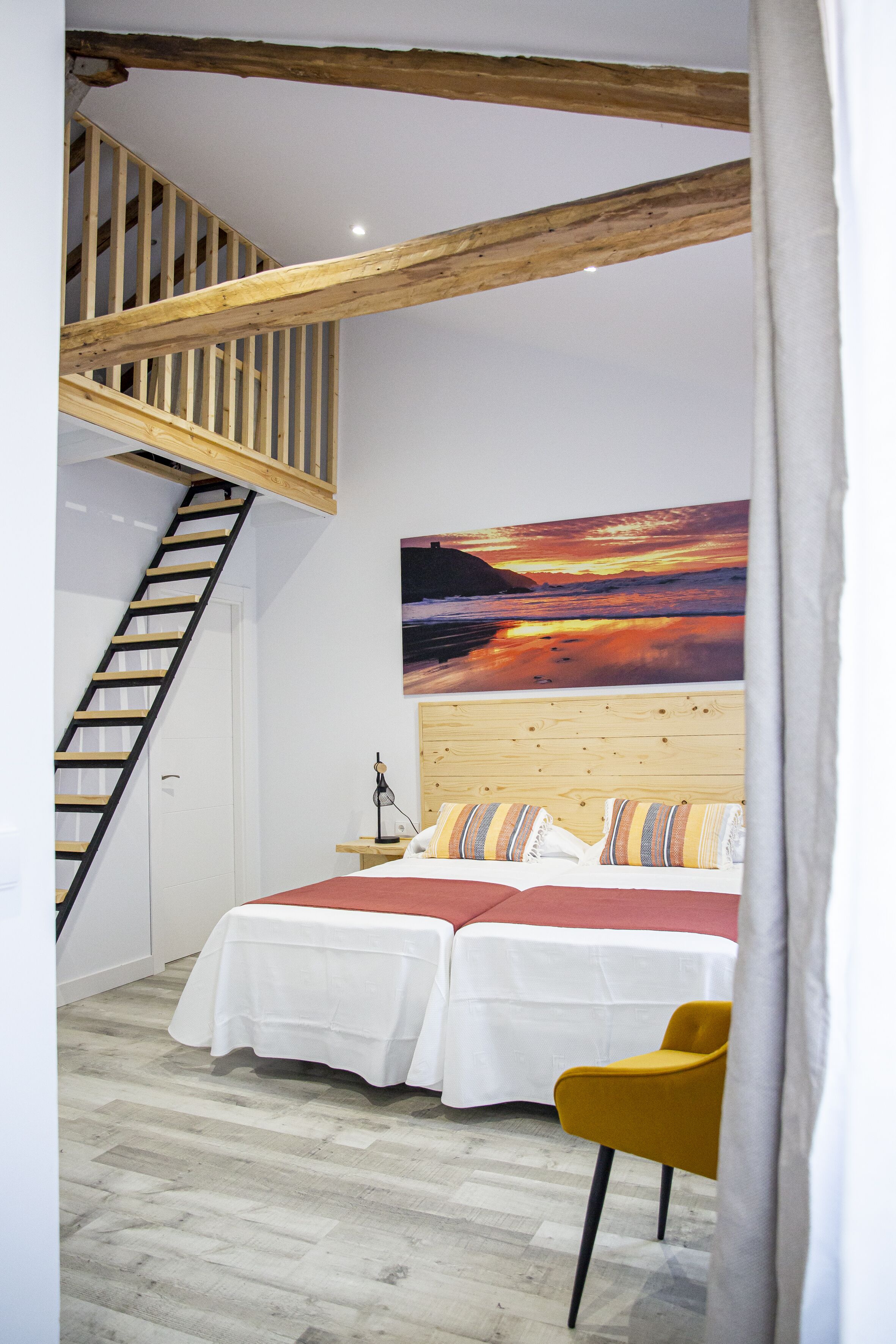 Un dormitorio con escalera de madera, cama con ropa blanca, una silla amarilla y un cuadro grande de un atardecer en la pared.
