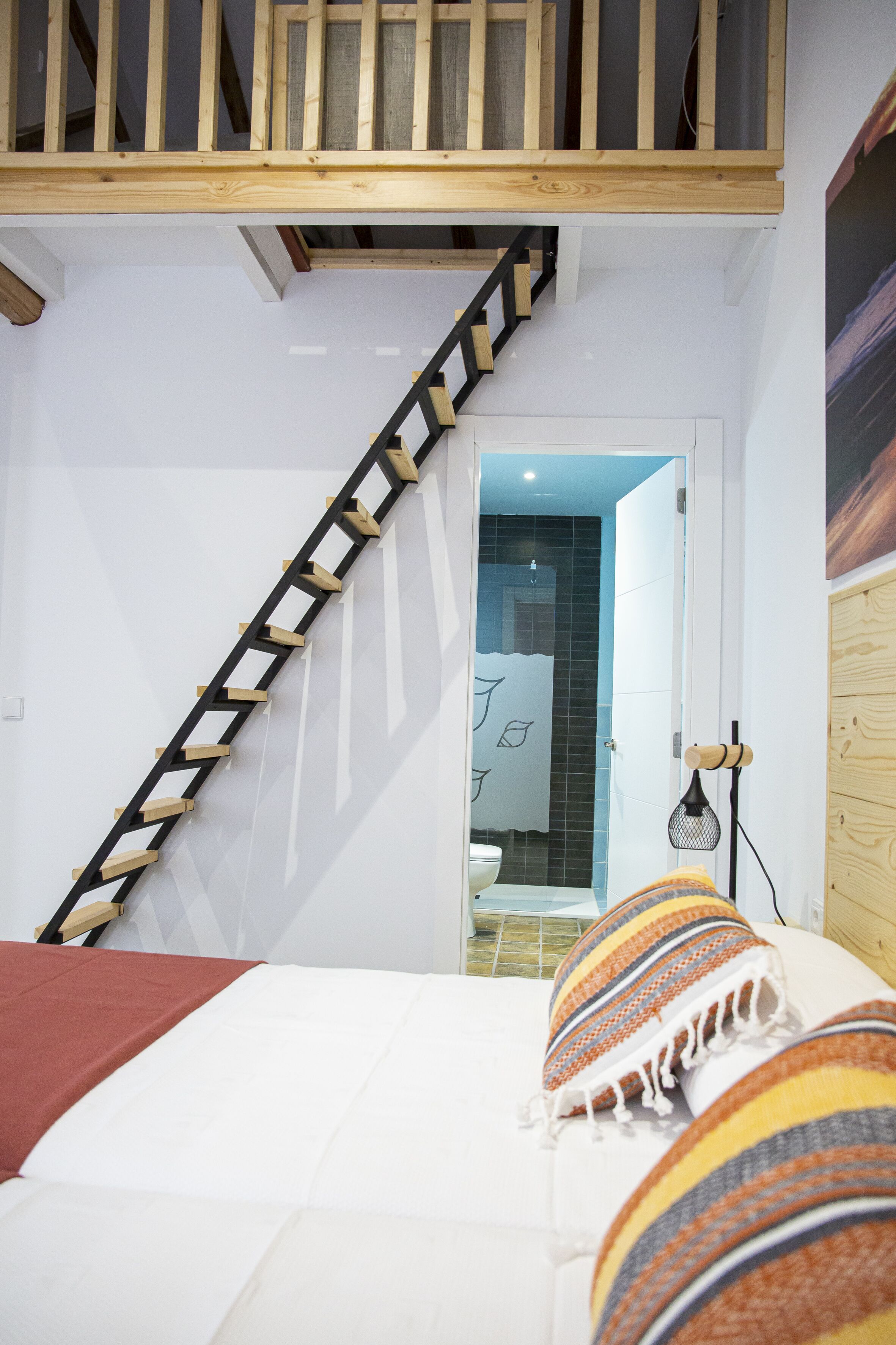 Un dormitorio con una escalera de madera, una cama con almohadillas estampadas y un baño visible a través de una puerta abierta.
