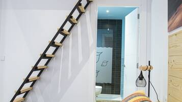 Un dormitorio con una escalera de madera, una cama con almohadillas estampadas y un baño visible a través de una puerta abierta.