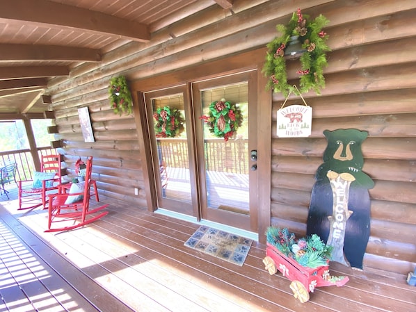A log cabin with a porch, two doors with wreaths, a welcome sign, a bear decoration, and red rocking chairs.
