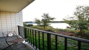 En balkon med metalstol og gelænder, med udsigt over vand og grøn bevoksning.