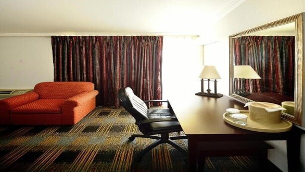 Une chambre d’hôtel avec un canapé rouge, un fauteuil de bureau noir, un bureau en bois avec une lampe, un plateau avec une tasse et une assiette, et un grand miroir au mur.