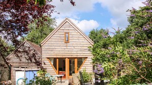Une cabane en bois avec un toit triangulaire, une véranda à l’avant et un jardin où poussent diverses plantes et fleurs.