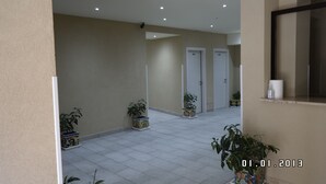 Un pasillo con suelo de baldosas, paredes color beige y puertas blancas. Hay plantas en macetas a lo largo de las paredes.
