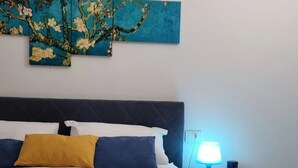 Una cama bien hecha con ropa de cama blanca y azul, un cojín amarillo y una lámpara azul en una mesita de noche.