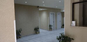 Un couloir au sol carrelé, aux murs de couleur beige et aux portes blanches. Des plantes en pot sont disposées le long des murs.