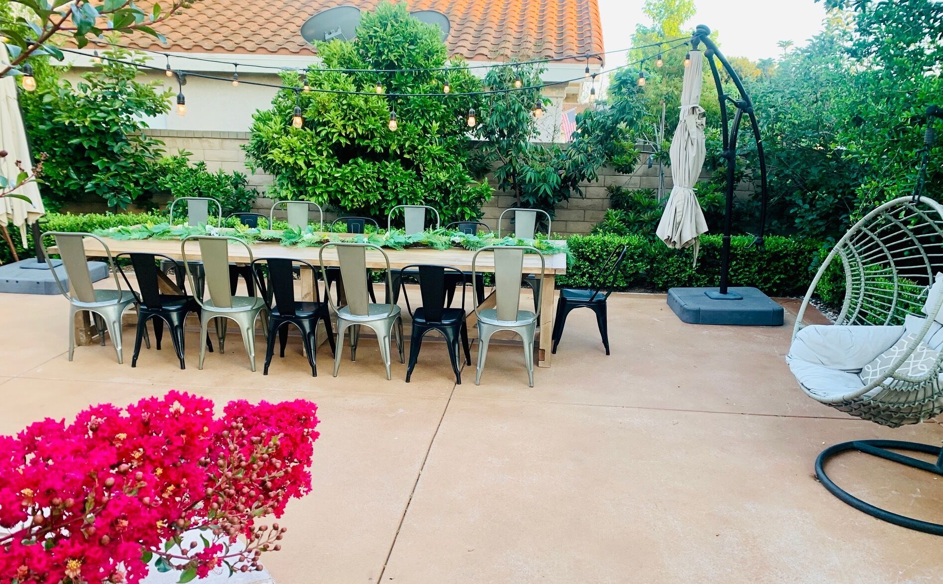 An outdoor patio with a wooden table and chairs, surrounded by greenery and a hanging chair.