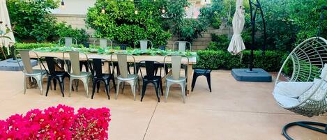 An outdoor patio with a wooden table and chairs, surrounded by greenery and a hanging chair.
