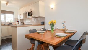 A modern kitchen with white cabinets, a microwave, and a dining area with a wooden table and chairs. - 7 Europa Court (Newquay)