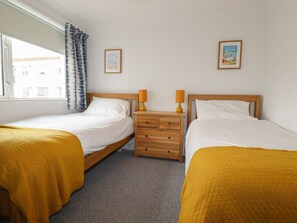 A double room with two single beds, a wooden nightstand with drawers, a lamp, and a window with a view of buildings. - 7 Europa Court (Newquay)