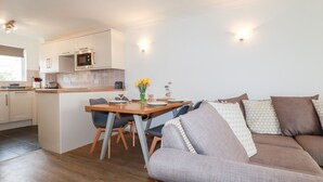 A modern kitchen with white cabinets, a microwave, and a dining area with a wooden table and chairs. - 7 Europa Court (Newquay)