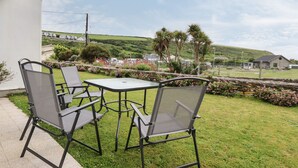 An outdoor patio with two chairs and a table, overlooking a grassy area with flowers and a distant view of houses and hills. - 7 Europa Court (Newquay)