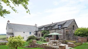 A stone house with a white exterior, a patio with a table and chairs, and a garden with a tree and flowers. - Honey Bee Cottage (Holsworthy)