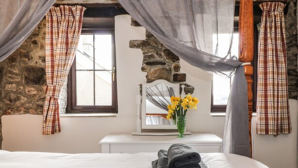 A bedroom with a stone wall, a bed with white linens, a dresser with a mirror, and a vase with yellow flowers. - Honey Bee Cottage (Holsworthy)