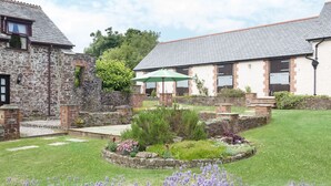 A courtyard with a stone wall, a grassy area, and purple flowers. - Honey Bee Cottage (Holsworthy)