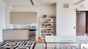 A modern kitchen with a microwave, sink, and cabinets. There's a bookshelf filled with books and a dining area with a table and chairs. An exit sign and a wheelchair accessibility sign are visible.