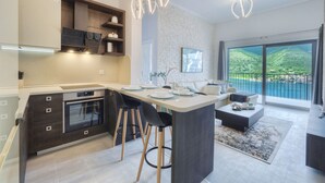 A modern kitchen with a central island, bar stools, and a large window offering a view of a mountainous landscape. - Luxurious Apartment with Captivating Majestic View (Dobrota)