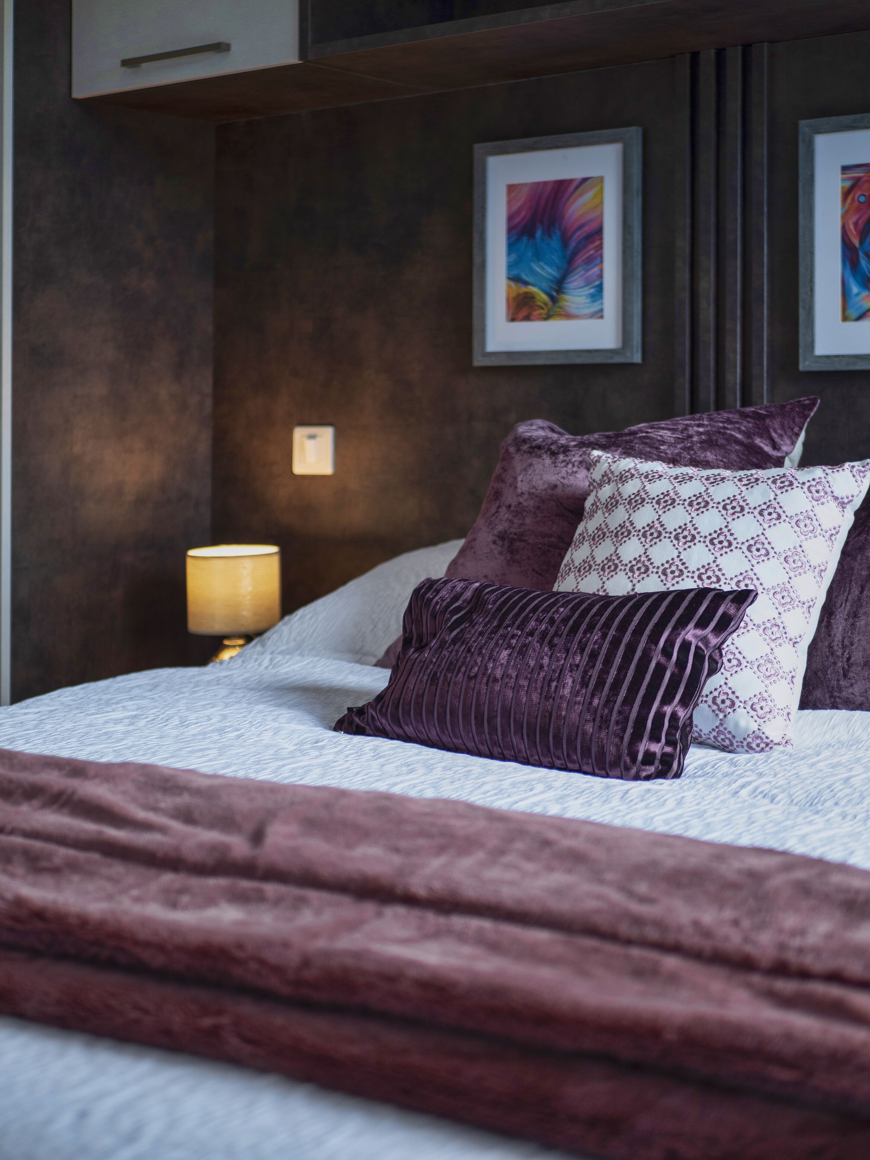 A neatly made bed with a dark purple blanket and pillows, a bedside lamp, and two framed artworks on the wall.