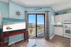 Un salon avec un téléviseur diffusant une scène de plage, un balcon avec vue sur la mer et un coin cuisine.
