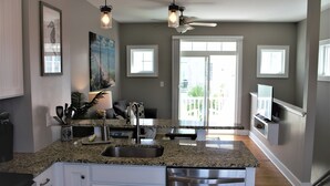 A modern kitchen with granite countertops, stainless steel appliances, and a large island with a sink. There are two pendant lights, a ceiling fan, and a large window with a view of the outdoors.