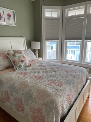 Une chambre à coucher avec un lit, des coussins à motifs floraux, une table de chevet avec une lampe, et une vue sur un balcon à travers une fenêtre munie de stores.