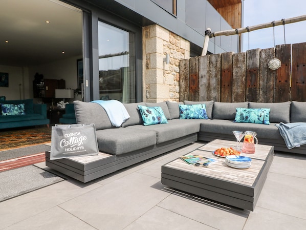 A modern outdoor seating area with a grey sectional sofa, a wooden coffee table, and a sign for Cornish Cottage Holidays.