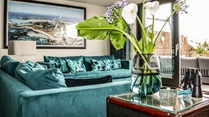 A teal sofa with patterned cushions, a glass coffee table with a vase of flowers, and a large framed landscape painting on the wall.