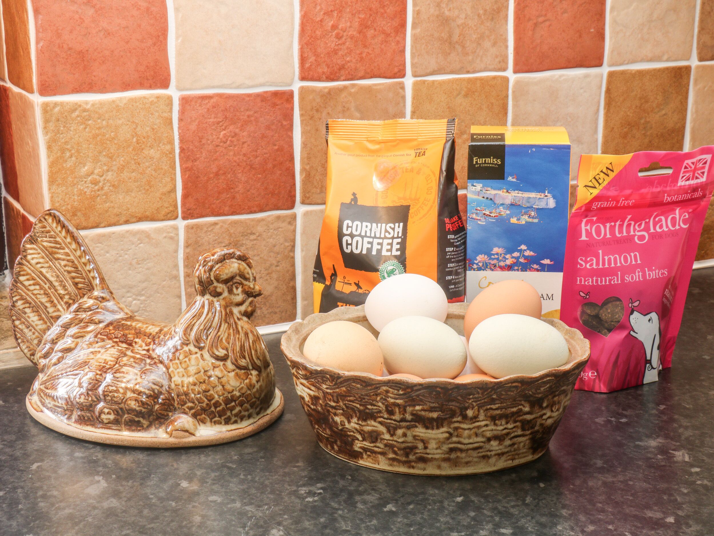 A kitchen counter with a ceramic rooster, a bowl of eggs, and various food products including coffee and dog food.