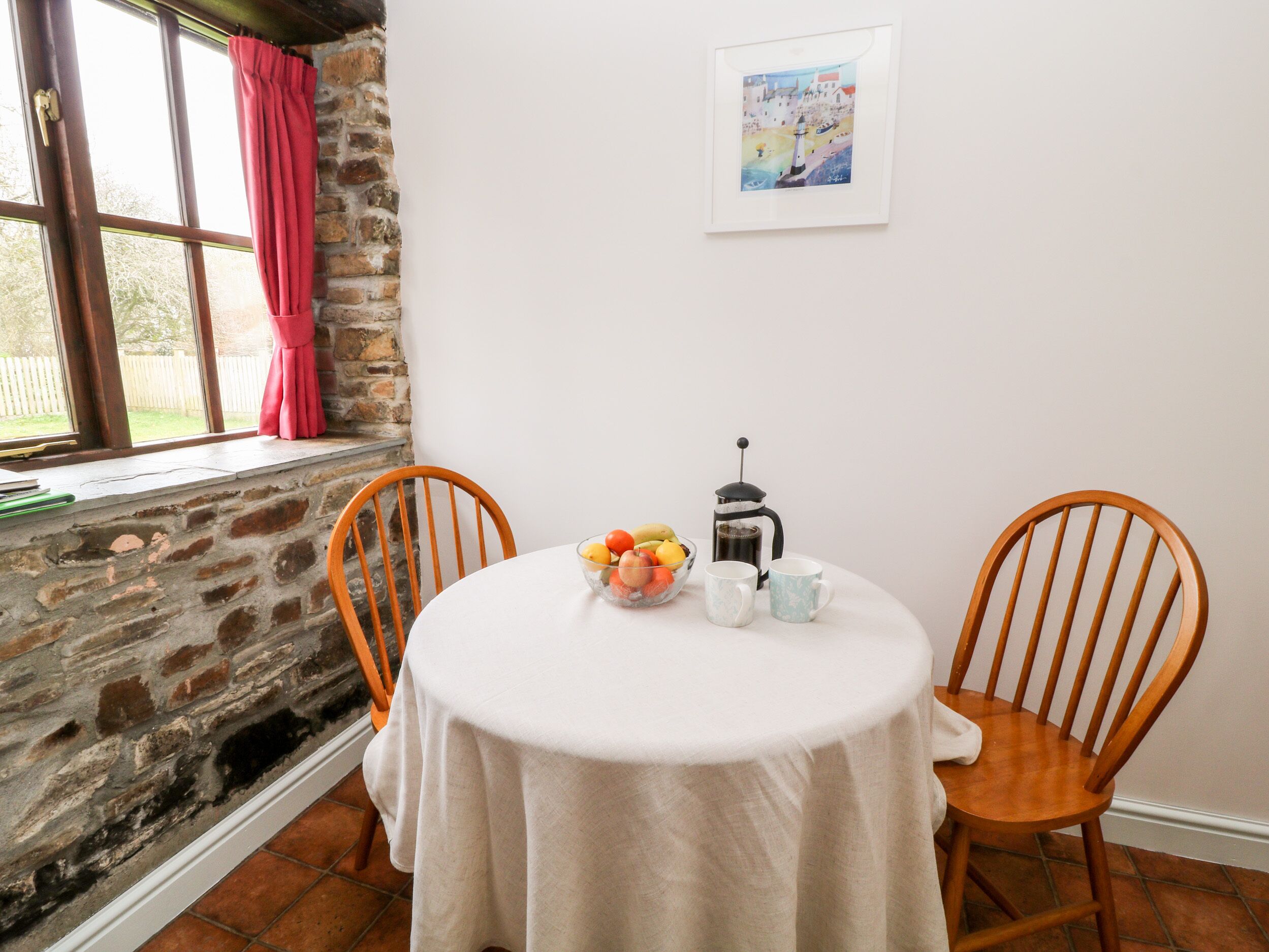 A dining area with a round table, two wooden chairs, a bowl of fruit, a coffee pot, and a framed picture on the wall.