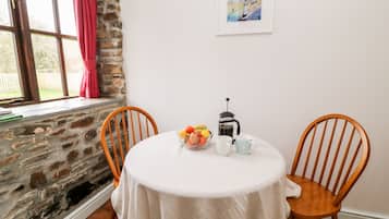 Un comedor con una mesa redonda, dos sillas de madera, un cuenco de fruta, una cafetera y un cuadro en la pared.