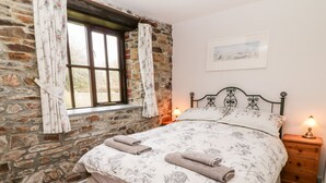 A bedroom with a stone wall, a bed with floral bedding, two bedside lamps, and a framed picture on the wall. - Wagtail (Holsworthy)