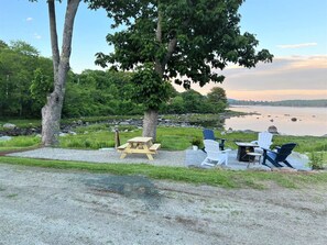 A lakeside patio with a picnic table, Adirondack chairs, and a small table with chairs.