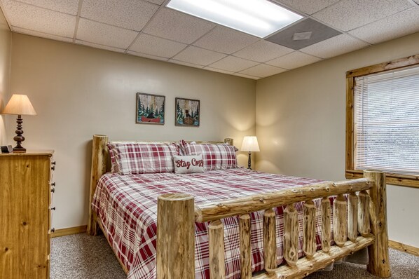 A bedroom with a wooden bed frame, a plaid bedspread, two wall-mounted paintings, a nightstand with a lamp, and a window with blinds. - 5 King BR/5 BA 🌟1 mile from PForge 🌟Sleeps 13 🌟Hot Tub (Pigeon Forge)